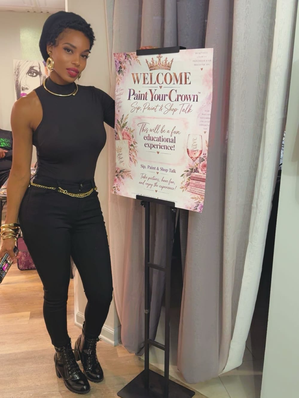 A guest posing beside the Paint Your Crown: Sip, Paint & Shop Talk welcome sign at Natural Touch Hair Studio